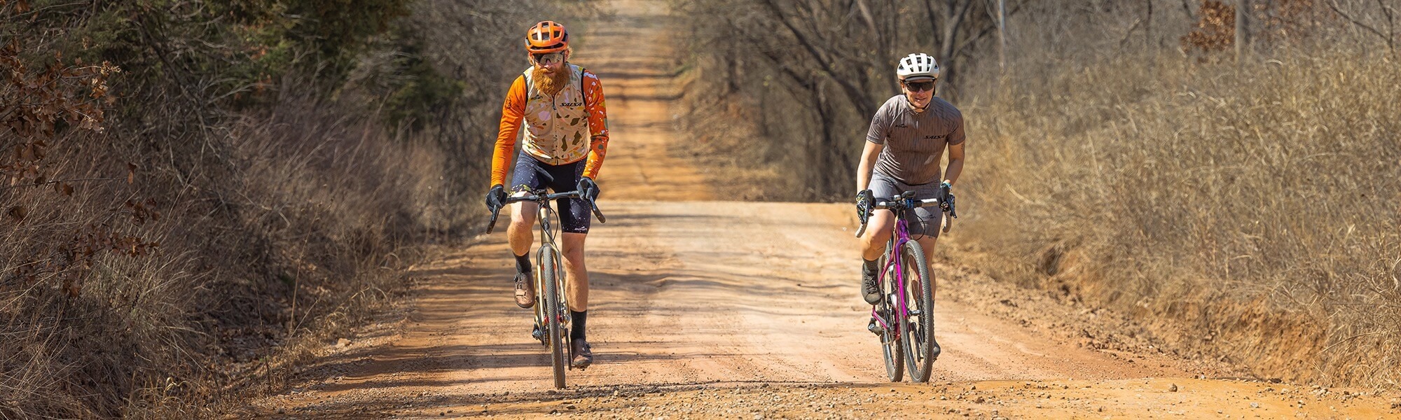 Two cyclists shown from the front ride drop bar bikes down a gravel road.